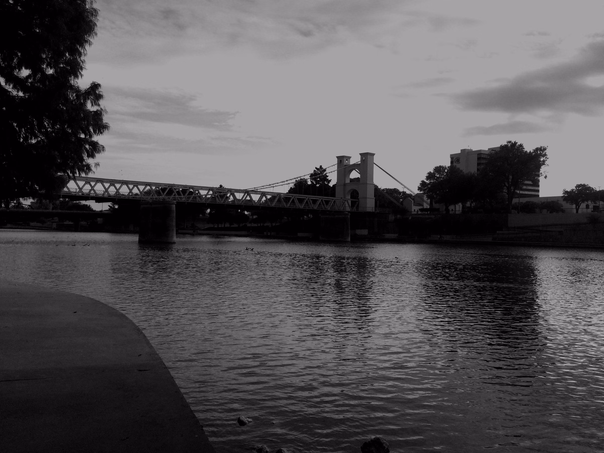 The Waco Suspension Bridge crossing the Brazos River in Waco, Texas, seen from the riverbank under a partly cloudy sky.