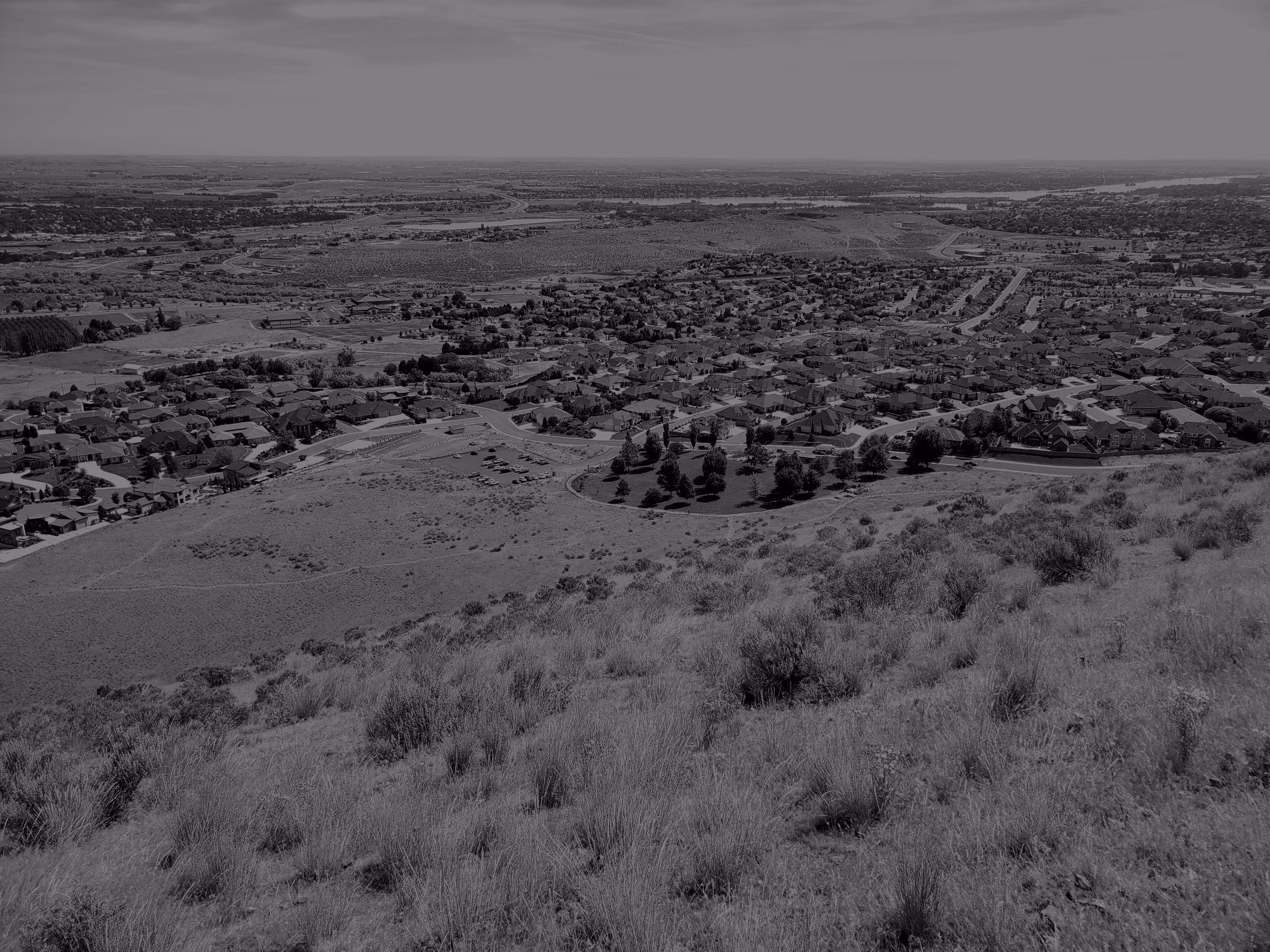 A view from a dry grassy hillside over the suburbs of Richland, Washington, with Trailhead Park in the foreground and flat country stretching to the horizon.