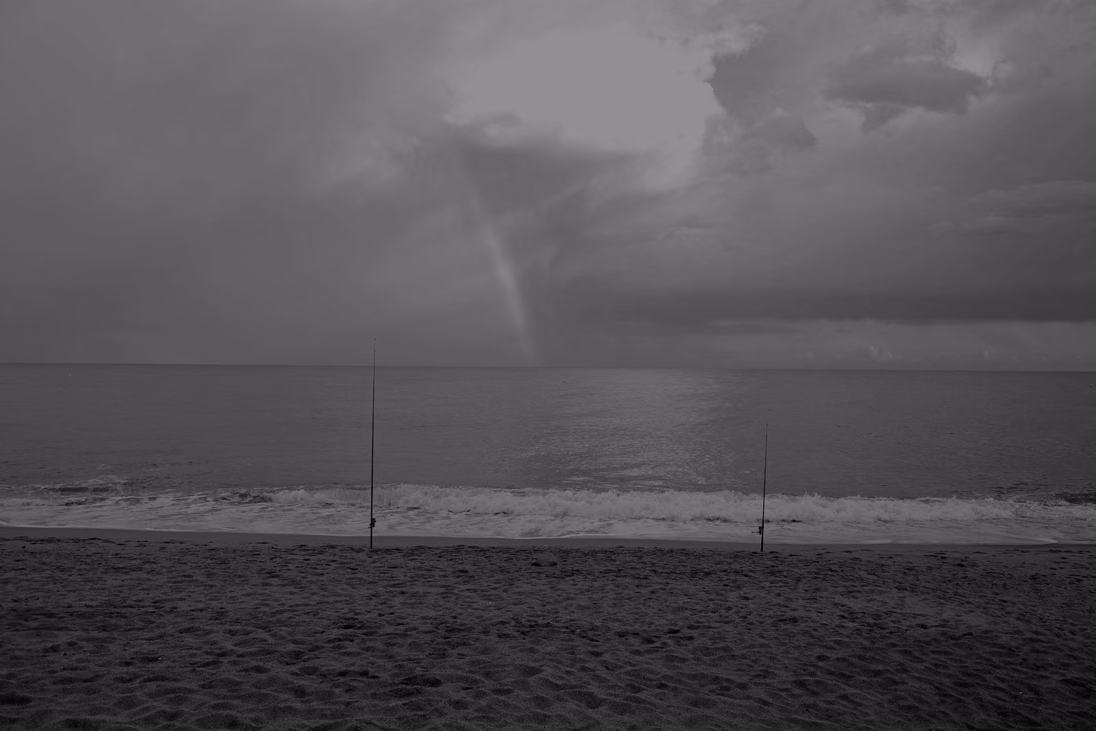 A quiet sandy beach at Pineda de Mar on the Costa del Maresme, with fishing rods planted in the sand and a rainbow under a cloudy sky.