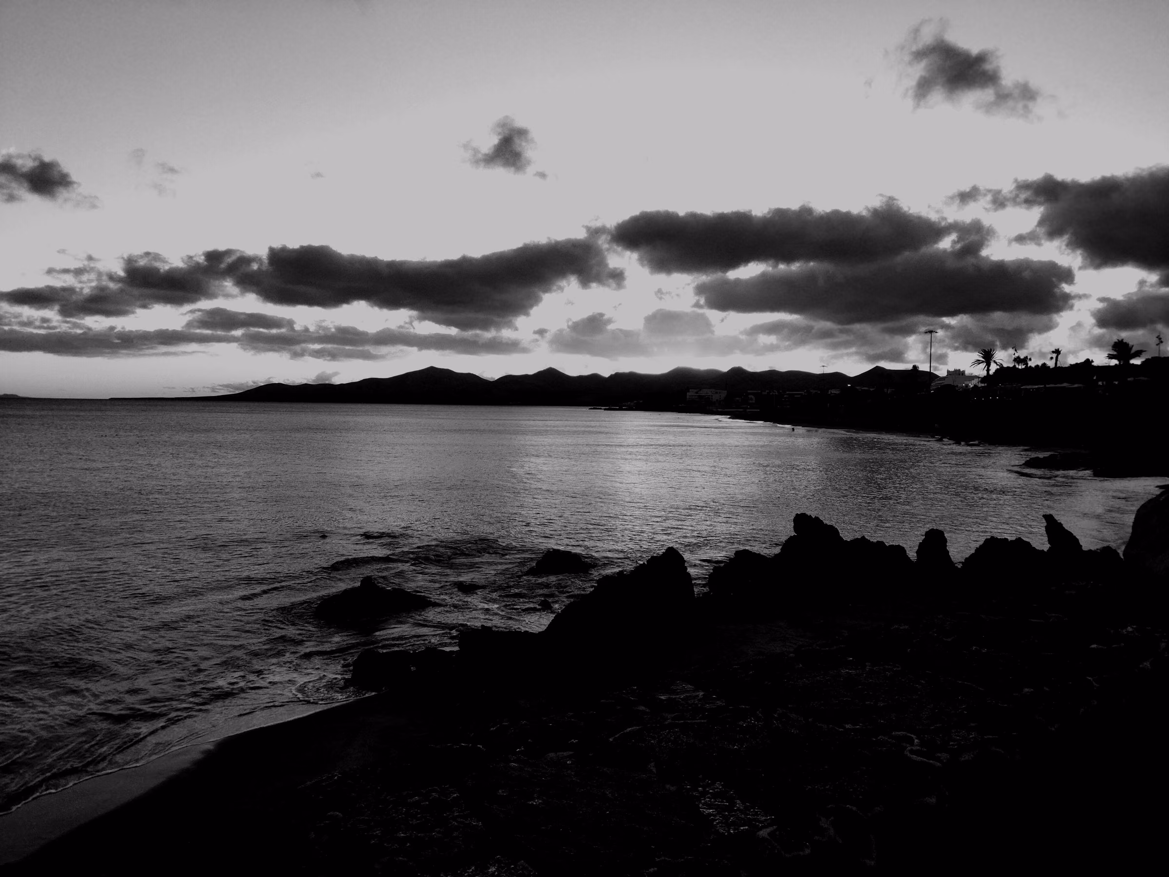 Dark volcanic rock above the bay at Playa Grande, Puerto del Carmen, with Lanzarote's hills on the horizon at dusk.